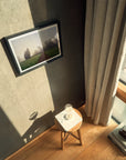 The image shows an interior space with a framed photograph of a foggy landscape hanging on a wall. The photo captures a misty morning in Surrey, England, with trees and a path visible. Sunlight streams through a window with beige curtains, casting shadows on the wall and floor. A wooden stool with a book and a glass sphere is placed near the window, adding a touch of elegance to the scene.